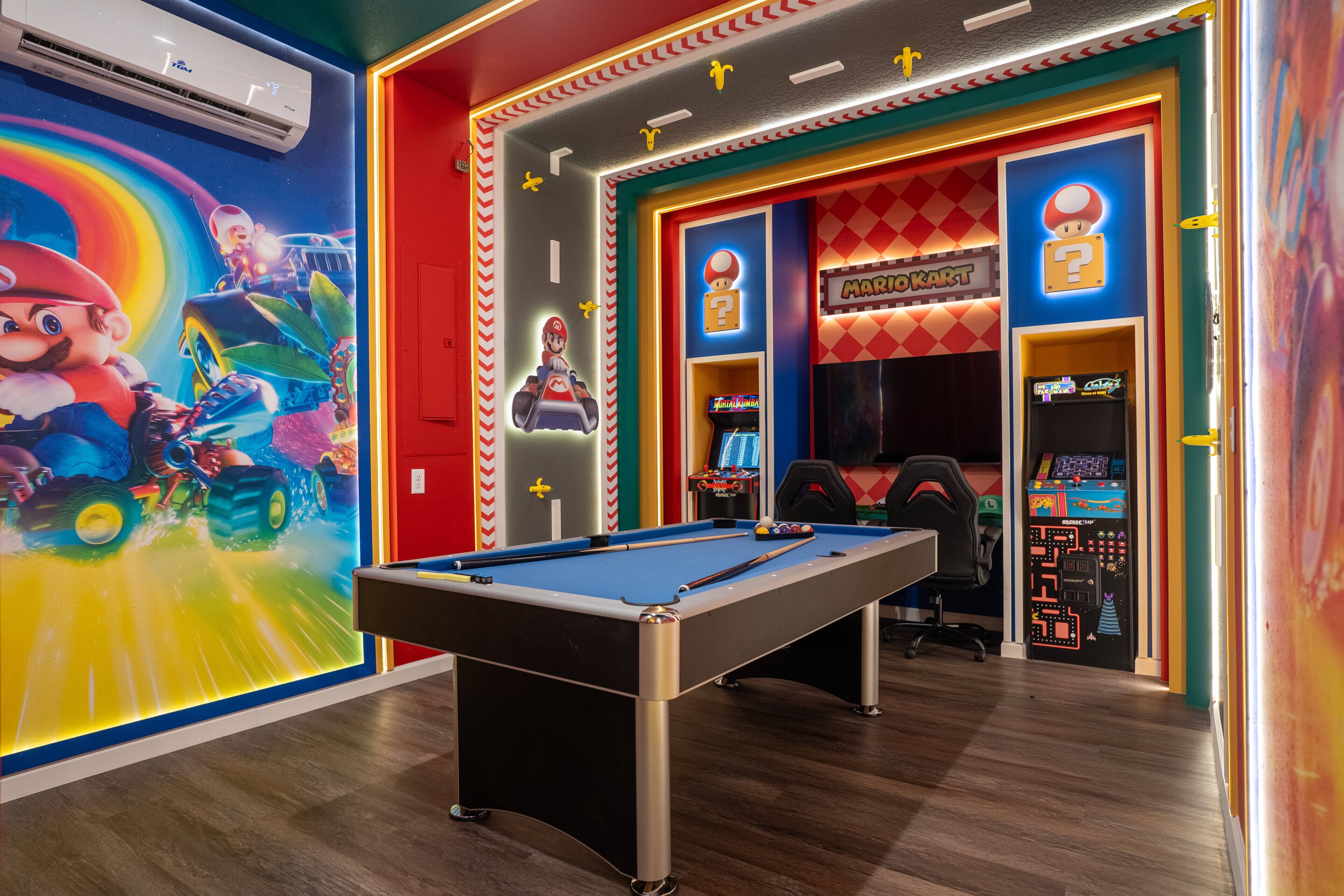 A pool table in a brightly decorated room with a Mario Kart arcade and a Mario-themed mural.