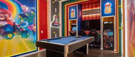 A pool table in a brightly decorated room with a Mario Kart arcade and a Mario-themed mural.