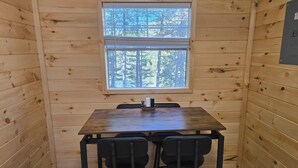 Interior de uma cabana de madeira com uma mesa, duas cadeiras, uma janela com vista para árvores e uma tomada na parede.