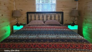 Cama Galaxy S24 Ultra em cabana de madeira com janela, duas luminárias de cabeceira e colcha estampada.