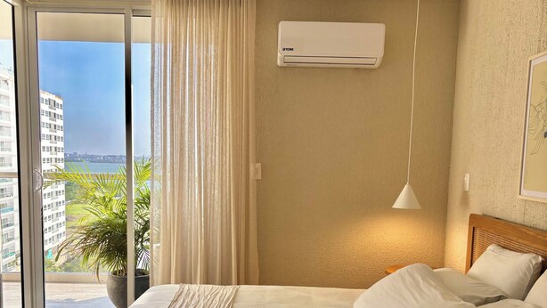 Ein Hotelzimmer mit einem großen Bett, einer Zimmerpflanze, Blick durch das Fenster und einer Klimaanlage.