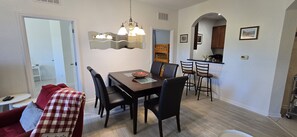 A dining area with a wooden table, black chairs, a red and white checkered sofa, a mirror, and a kitchen in the background. - Last Minute Disney Getaway! Luxury Condo with Private Balcony. Book Now! #173229 (Orlando)