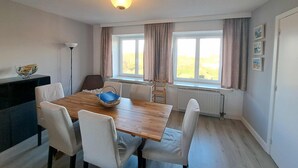 A dining area with a wooden table, white chairs, a basket, and a radiator under a window with curtains. - Spacious Family Stay in Oostduinkerke (Koksijde)