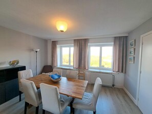 A dining area with a wooden table, white chairs, a basket, and a radiator under a window with curtains. - Spacious Family Stay in Oostduinkerke (Koksijde)