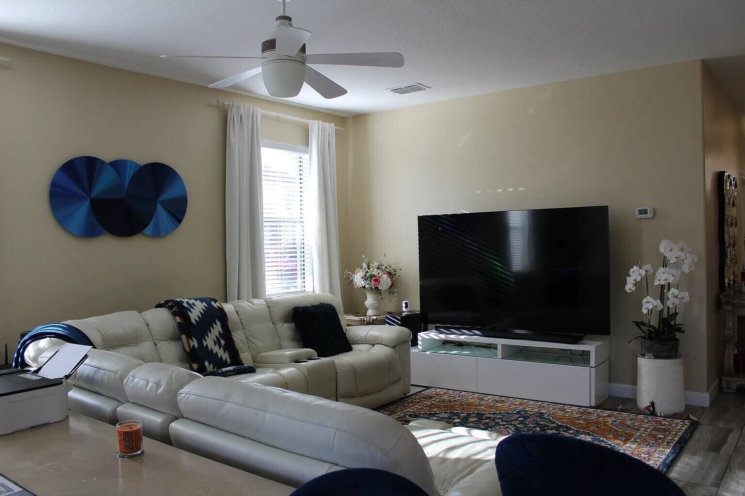 Un salon comprenant un canapé, un téléviseur à écran plat, un ventilateur de plafond et une fenêtre avec des rideaux.