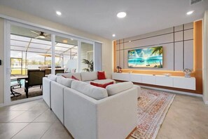 A modern living room with a white sofa, a TV mounted on the wall, and a glass door leading to an outdoor area. - Last Minute Disney Getaway! Private Pool & Themed Bedrooms. Book Now! #173535 (Orlando)