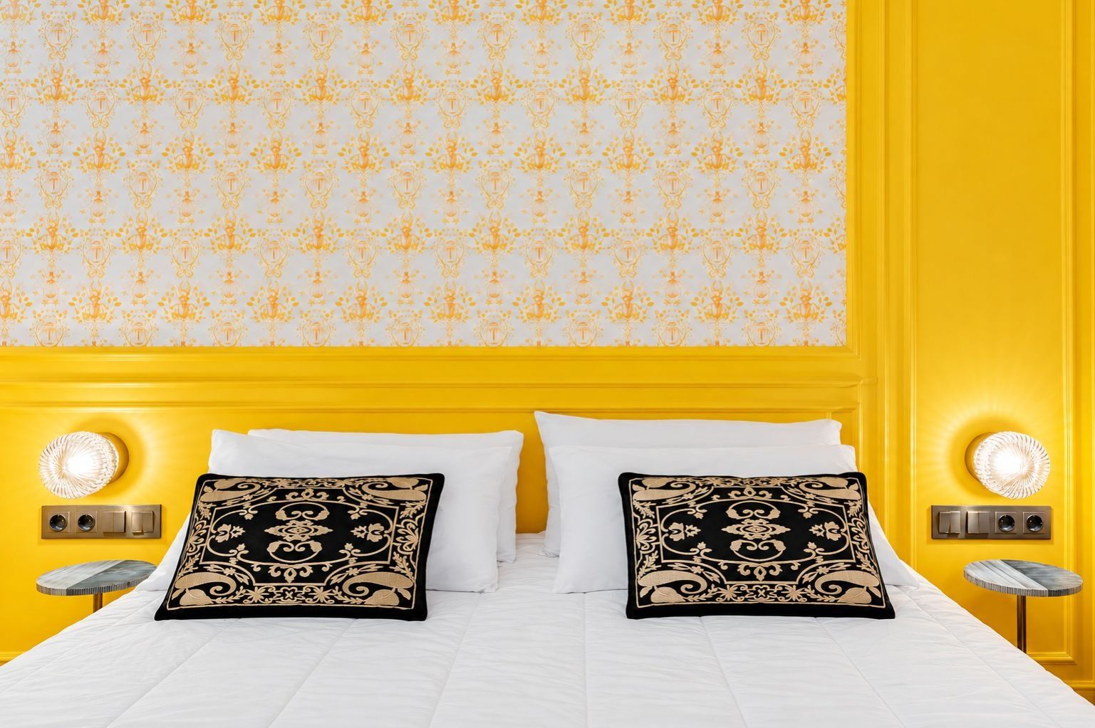 Een hotelkamer met een geel accentmuur, behang met een patroon en twee decoratieve kussens op een wit bed.