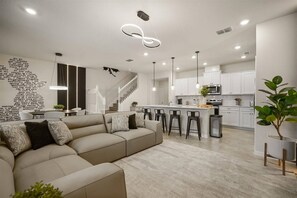 Ein modernes Wohnzimmer mit einer Couch, einer Essecke mit Barhocker, einer Küche mit weißen Schränken und einer Treppe.