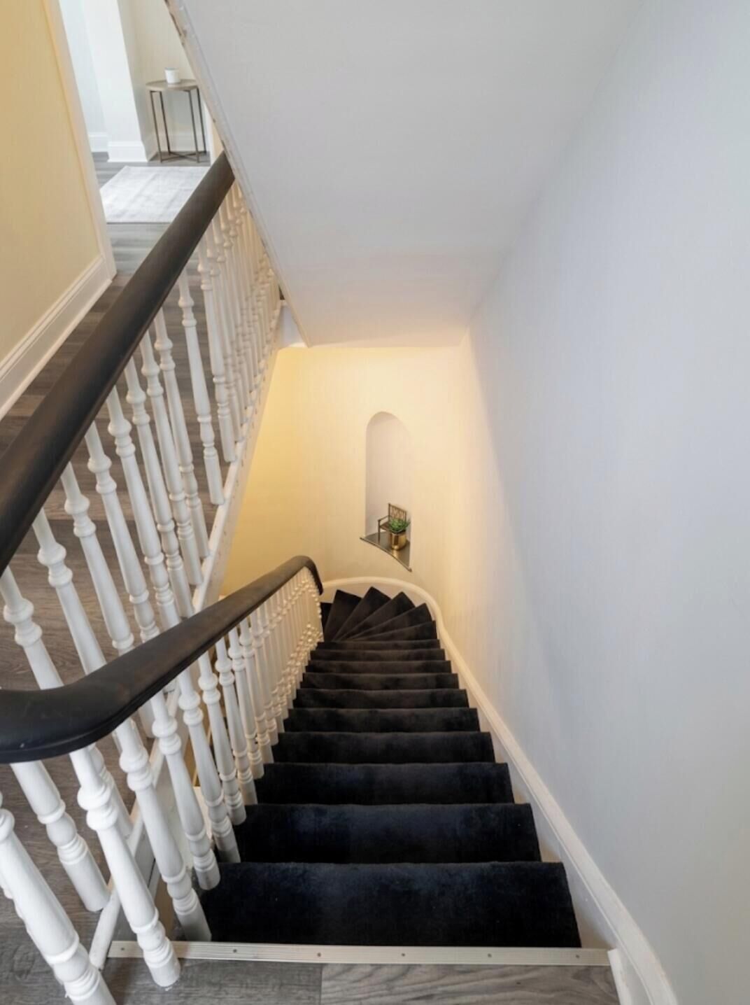 Un escalier avec des balustrades blanches et des rampes noires, menant à un palier où se trouvent une petite table et une plante en pot.