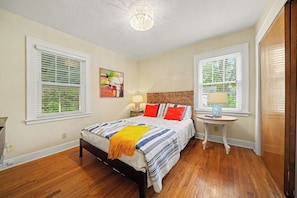 A bedroom with a bed, two bedside tables, a nightstand, a ceiling light, and two windows with blinds.
