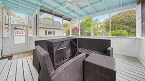 En inglasad veranda med möbler i rotting, en takfläkt och utsikt över ett hus och träd.
