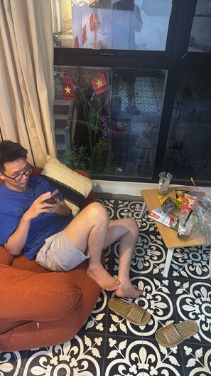 Eine Person sitzt auf einem Sofa, dabei hält sie ein Smartphone. Auf einem Tisch stehen Snacks und ein Getränk, und durch das Fenster weht eine Flagge.