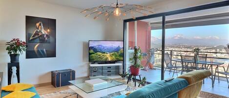 Una sala de estar moderna con un televisor grande, una lámpara de techo original, un sofá y vistas a montañas a través de amplios ventanales.