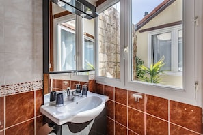 A bathroom with a white sink, two dark-colored mugs, a mirror, and a window with a view of a stone wall and greenery outside.