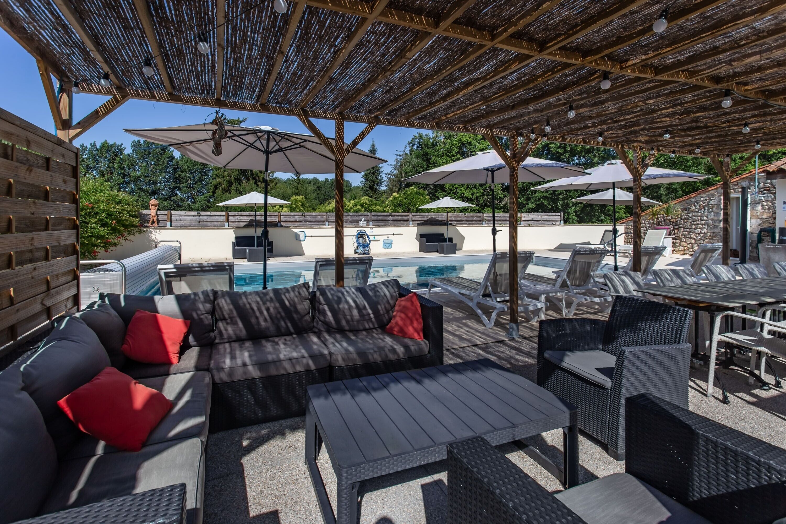 Un espace piscine extérieur avec des chaises longues et des parasols, un coin salon couvert avec des canapés et des tables, et un mur de pierre en arrière-plan.