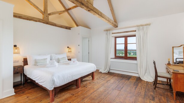 A bedroom with a wooden floor, a bed with white linens, a window with curtains, and a wooden dresser. - The Farmhouse at Nunwell (Brading)
