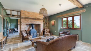A rustic living room with a brick fireplace, leather furniture, and a large window. - The Farmhouse at Nunwell (Brading)