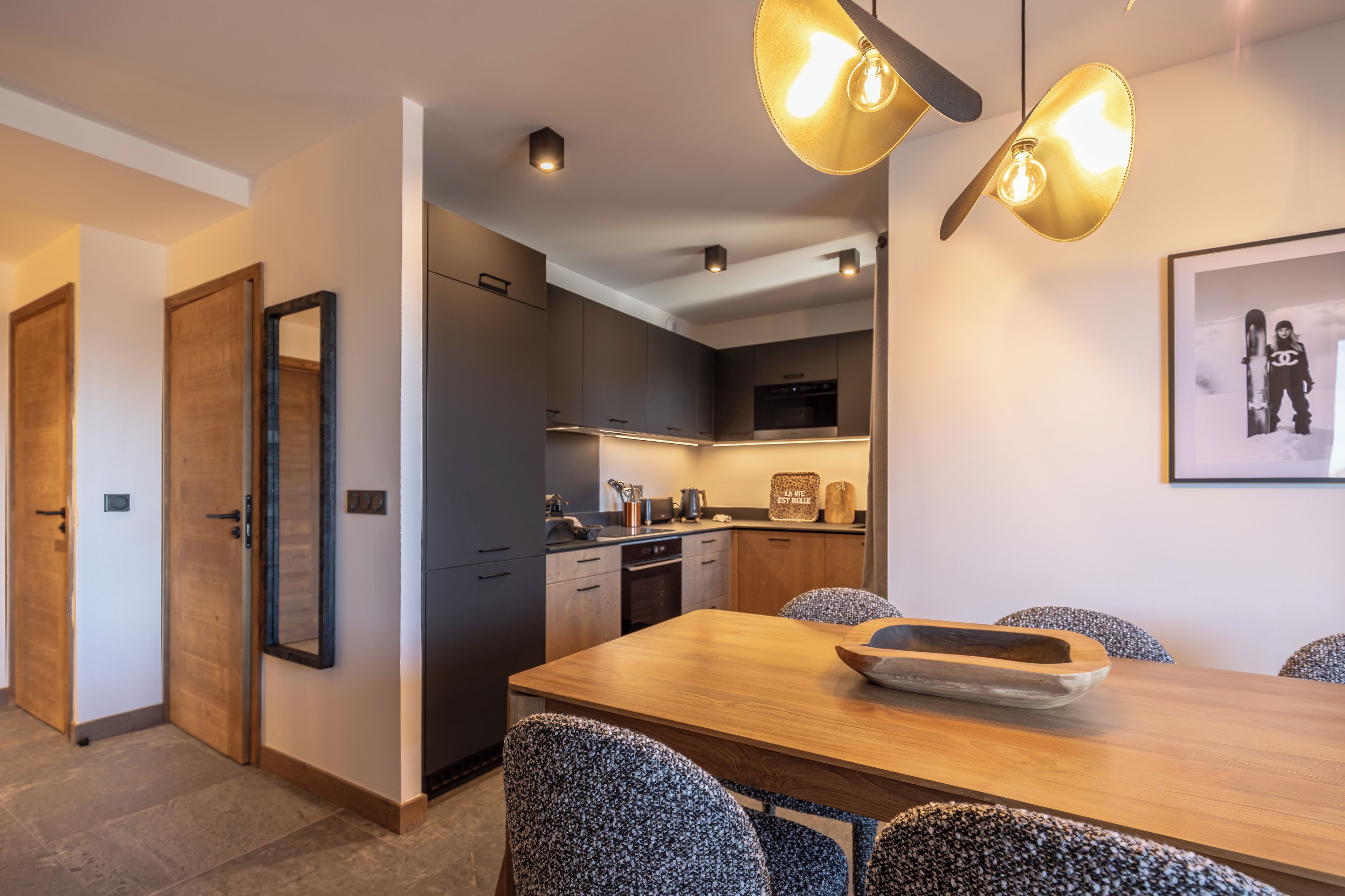 Cocina moderna con armarios oscuros y una isla central, un área de comedor con mesa de madera y sillas estampadas, y un cuadro enmarcado en la pared.