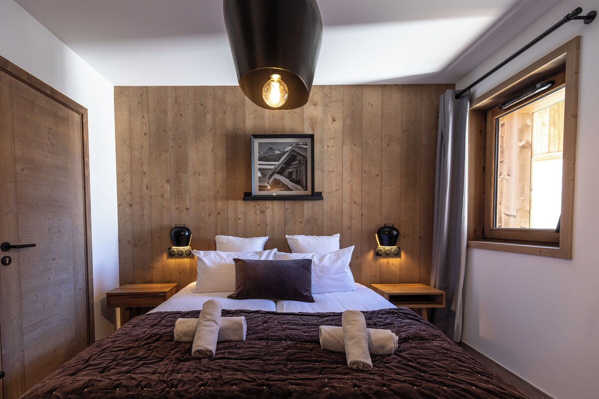 A bedroom with a wooden headboard, a bed with white and brown bedding, two bedside tables with lamps, and a window with curtains.
