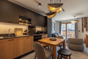 A modern kitchen with a built-in oven, a dining area with a wooden table and patterned chairs, and a view of the city through large windows. - TERRESENS - La Perle d'Alba (Huez)