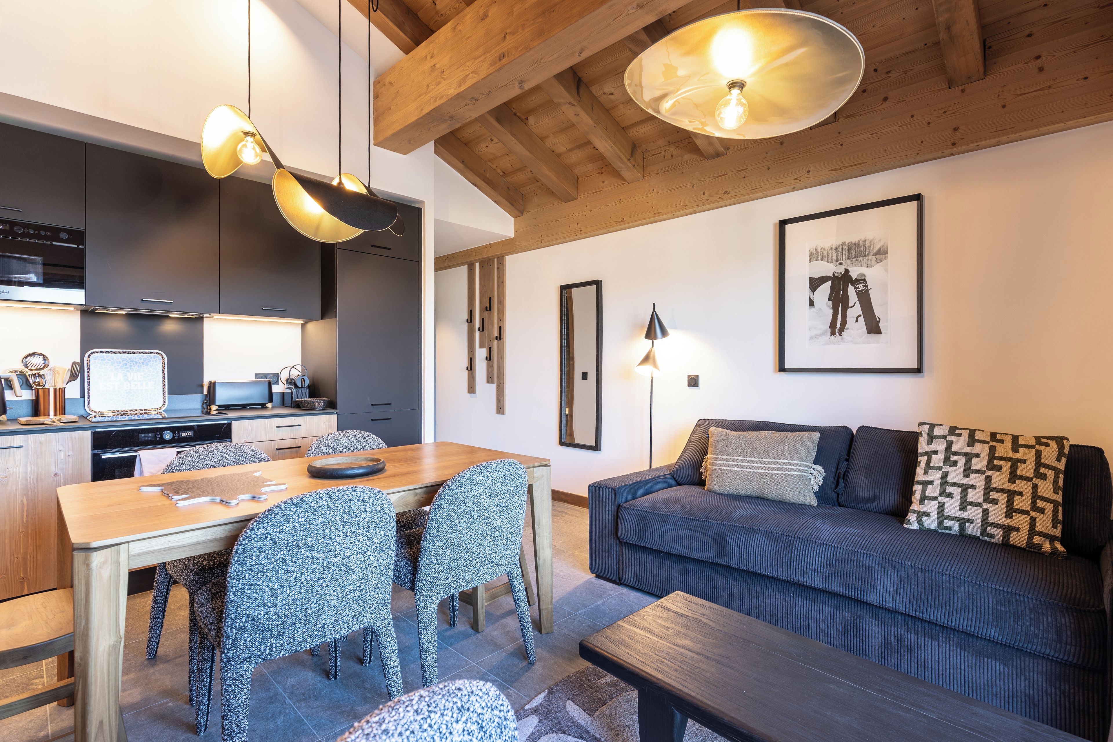 A modern kitchen with dark cabinetry, a dining table with chairs, and a wooden ceiling.