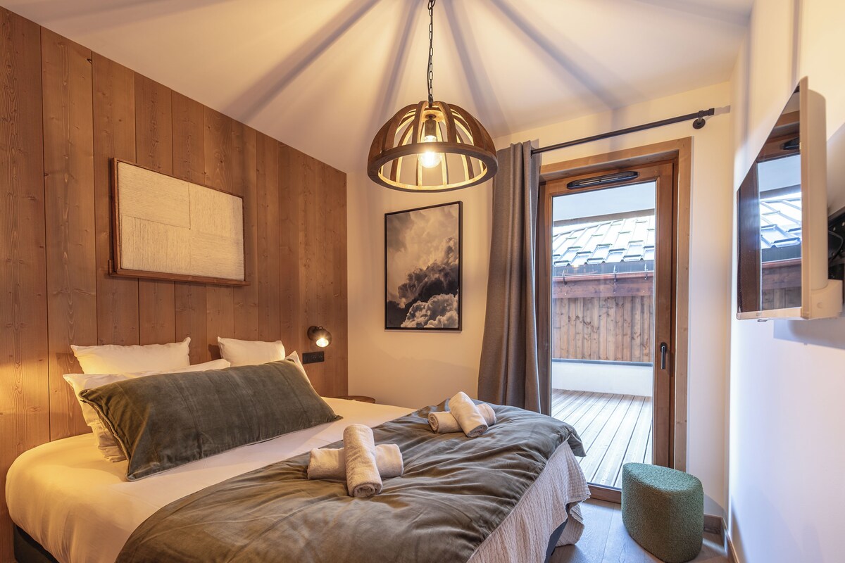 A bedroom with a wooden headboard, a bed with white and grey bedding, a mounted picture, a hanging light fixture, and a view of the outside through a window.