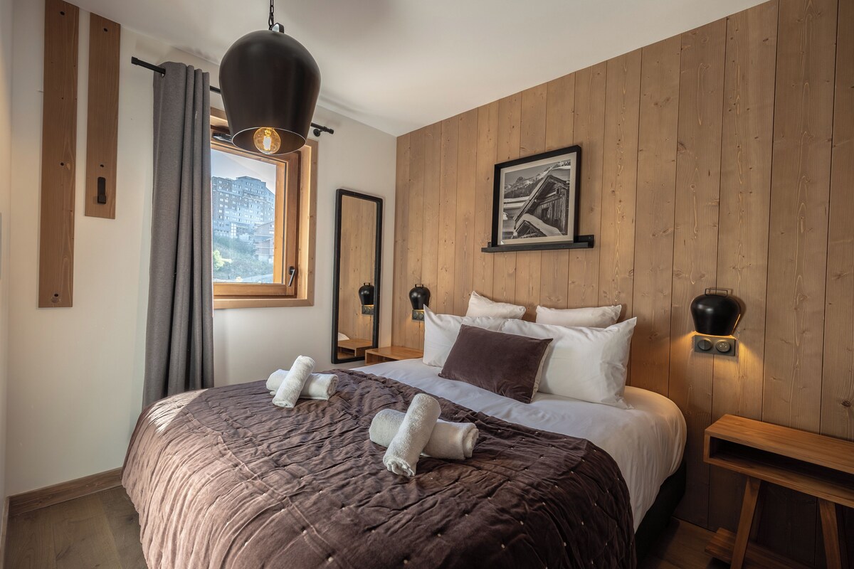 A bedroom with a large bed, wooden panel walls, a window with curtains, a framed picture, and bedside lamps.