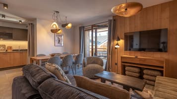 A modern living room with a sofa, dining table, and chairs, a kitchen area, and a television mounted on a wooden wall.