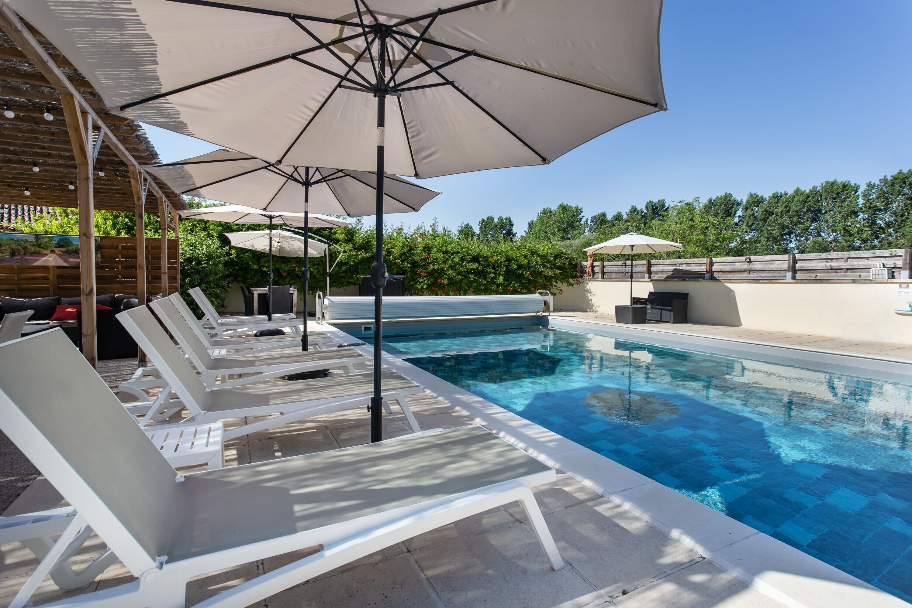 Una piscina con sillones de descanso y sombrillas, rodeada de una terraza de madera y follaje.