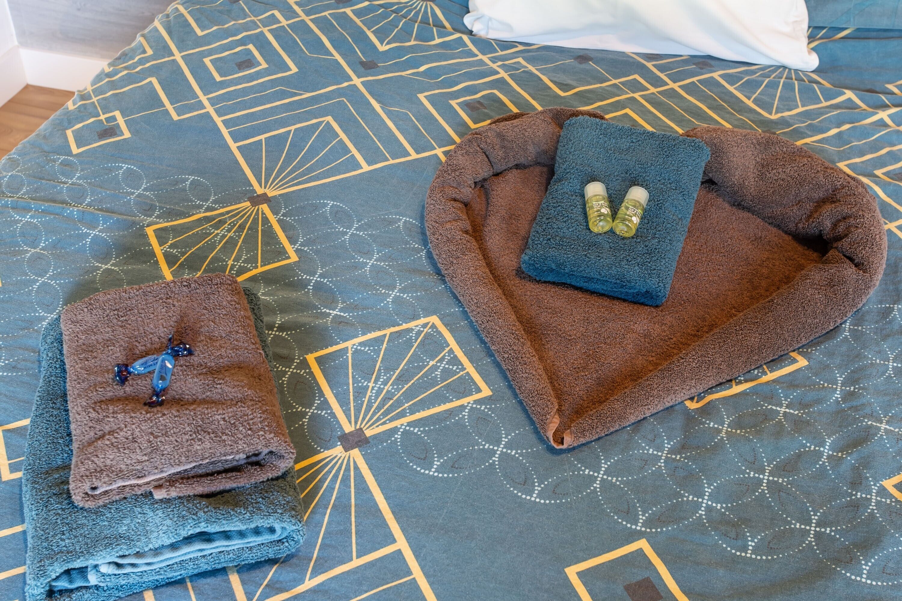 Uma cama com colcha estampada geometricamente, uma toalha marrom e azul, e duas garrafas sobre a cama.
