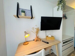Ein Zimmer mit Schreibtisch, Wand-Fernseher, Ablage mit Pflanze, Lampe und Fernbedienung.