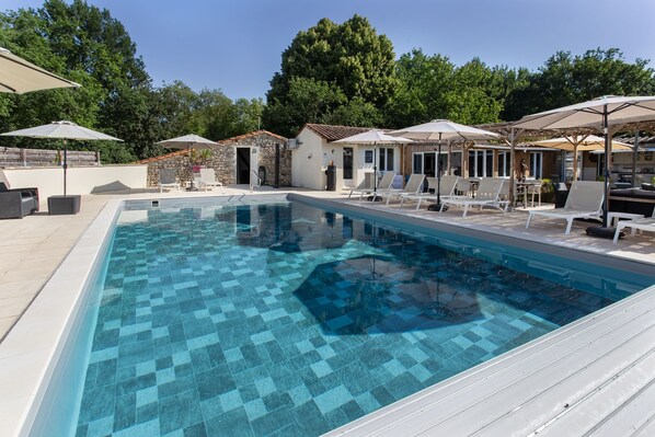 Une piscine avec un sol carrelé, des chaises longues, des parasols et un bâtiment en pierre en arrière-plan.