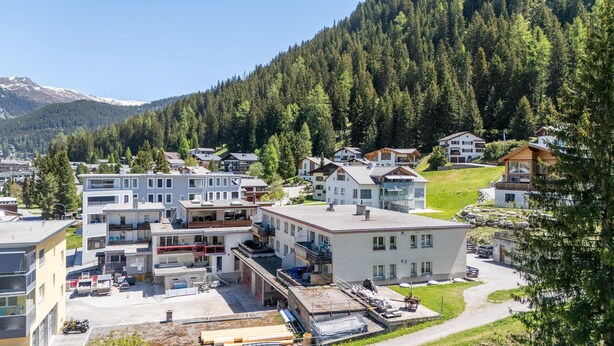 Una zona residenziale con case moderne, un cantiere edile in vista e montagne boscose sullo sfondo.