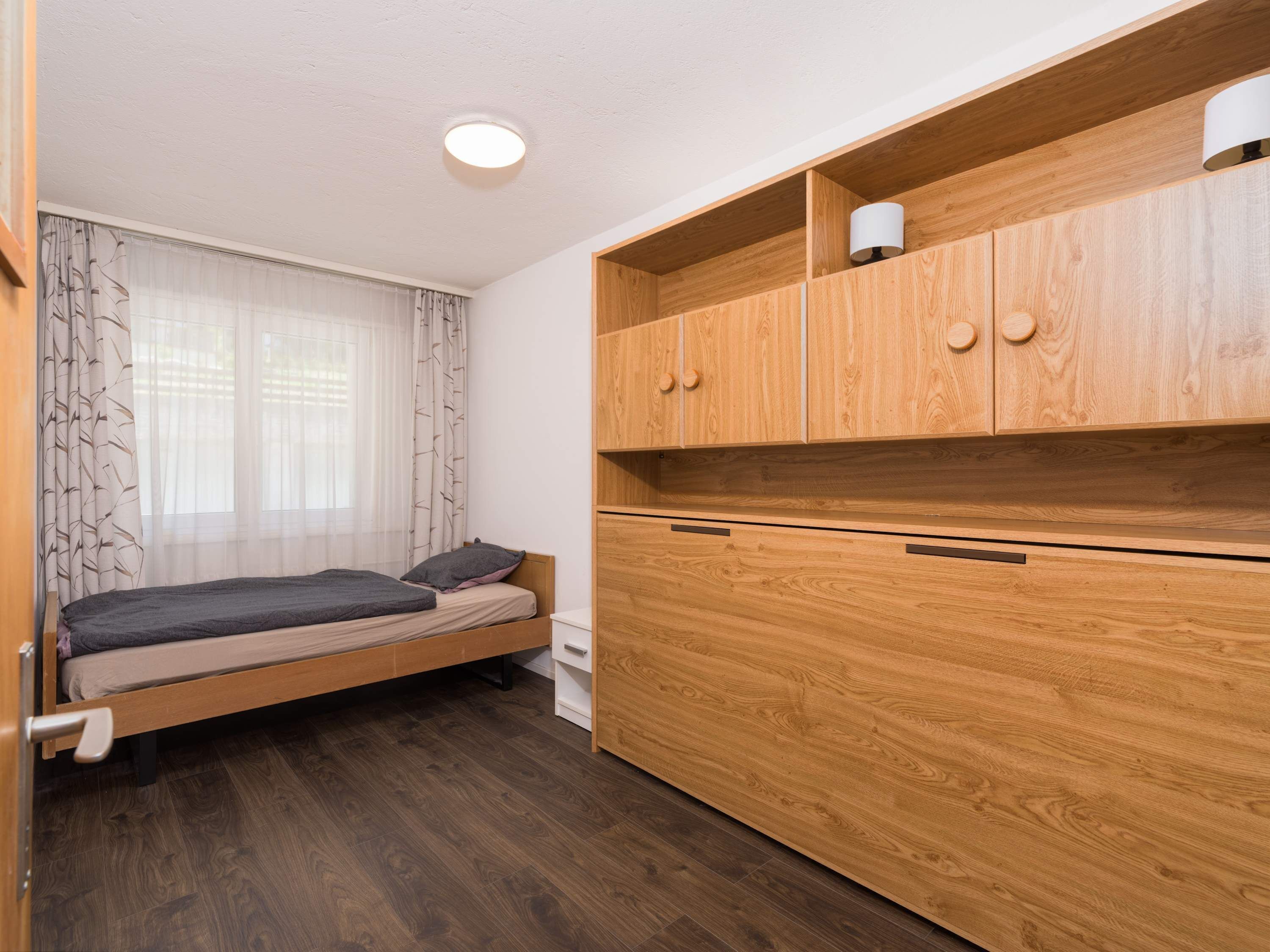 Ein kleines Zimmer mit Bett, Holzschrank und Fenster mit durchsichtigen Vorhängen.