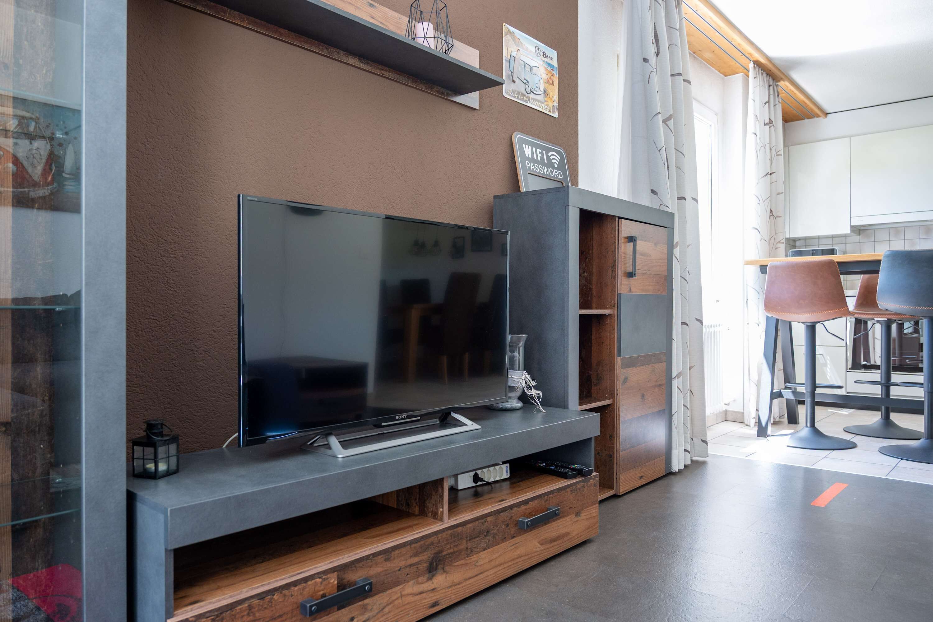 Una sala de estar moderna con un televisor de pantalla plana, almacenamiento de madera y una cocina al fondo.