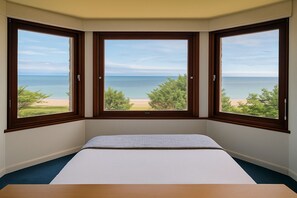 A bedroom with a large bay window overlooking a beach and ocean.