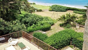 A balcony with a view of the beach, a sandy area with lounge chairs, and a fenced garden.