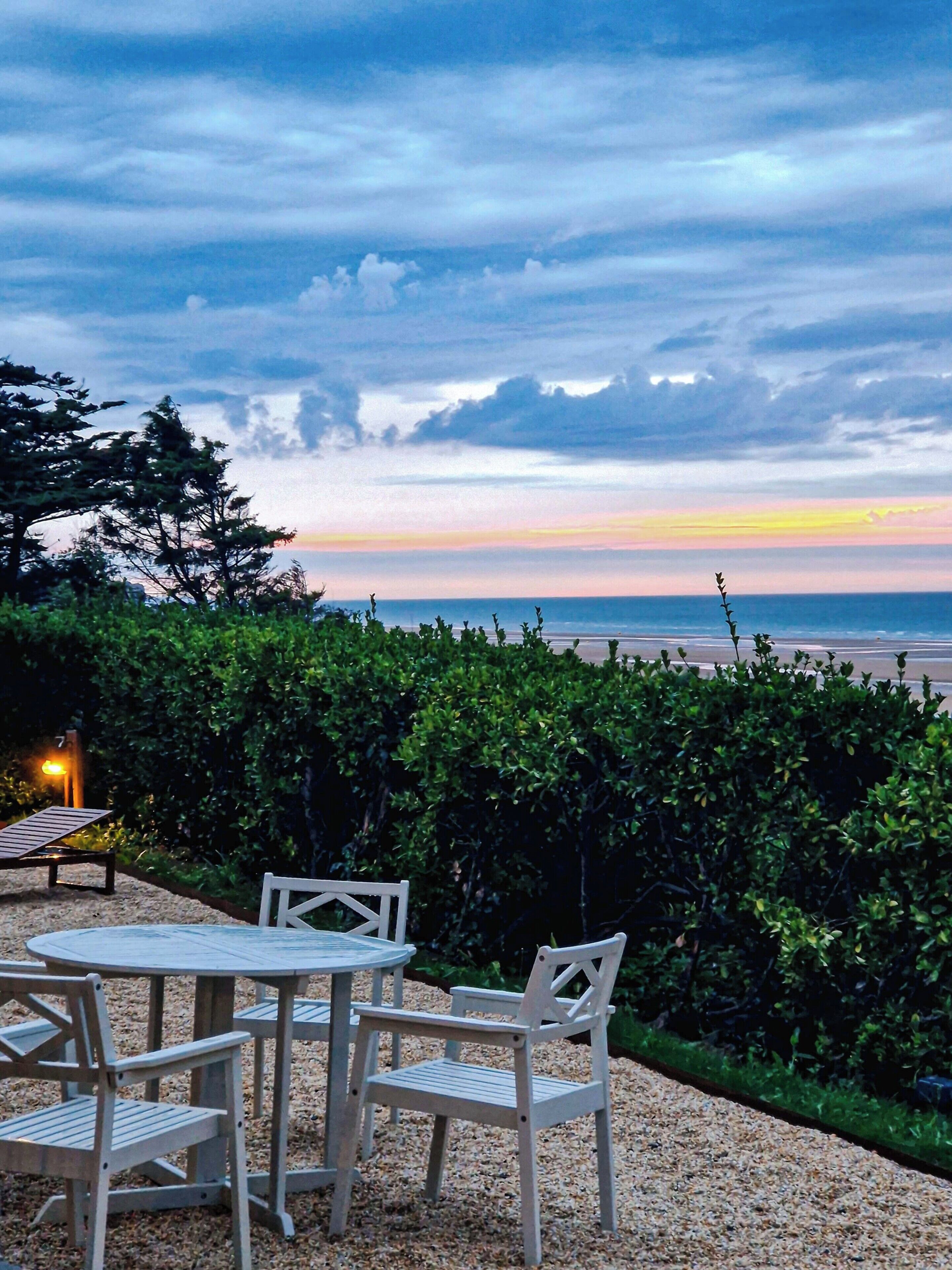 An outdoor seating area with a table and chairs, a hedge, and a view of the beach and ocean.