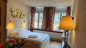 A hotel room with a bed, a desk, a lamp, a window with curtains, and a wall decoration. - Clemanbruges (Bruges)