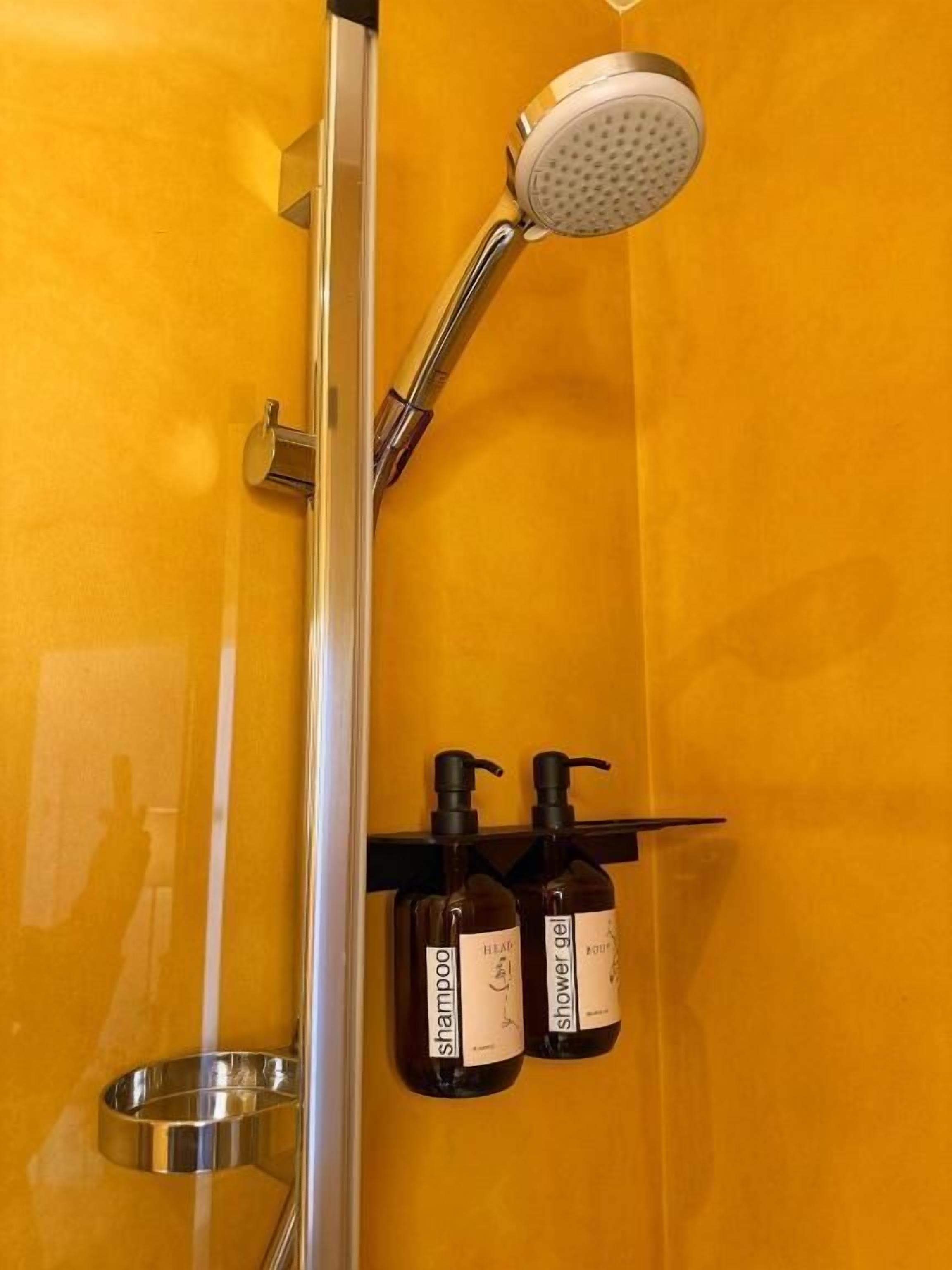 Ducha con cabezal de ducha manual, lavabo metálico y dos botellas de champú y gel de ducha sobre una pared amarilla.