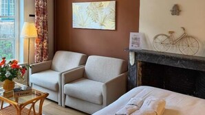 A hotel room with a bed, two armchairs, a table with a vase of flowers, a fireplace, and a city view through the window. - Clemanbruges (Bruges)