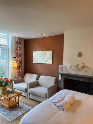 A hotel room with a bed, two armchairs, a small table with a lamp, a painting on the wall, a bicycle decoration, and a fireplace. - Clemanbruges (Bruges)