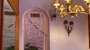 A staircase leading to a landing with a chandelier, a wall-mounted fire exit sign, and a mirror on the left. - Clemanbruges (Bruges)