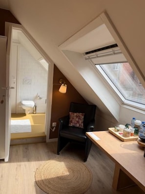 Una habitación pequeña y acogedora con tragaluces, una silla, una mesa con diversos objetos y vistas al exterior.