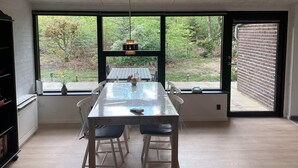 Uma sala de jantar com uma mesa grande e cadeiras, um lustre pendurado e vista para um jardim através de amplas janelas.