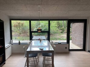 Ein Essbereich mit großem Tisch und Stühlen, einer hängenden Leuchte und Blick auf einen Garten durch große Fenster.