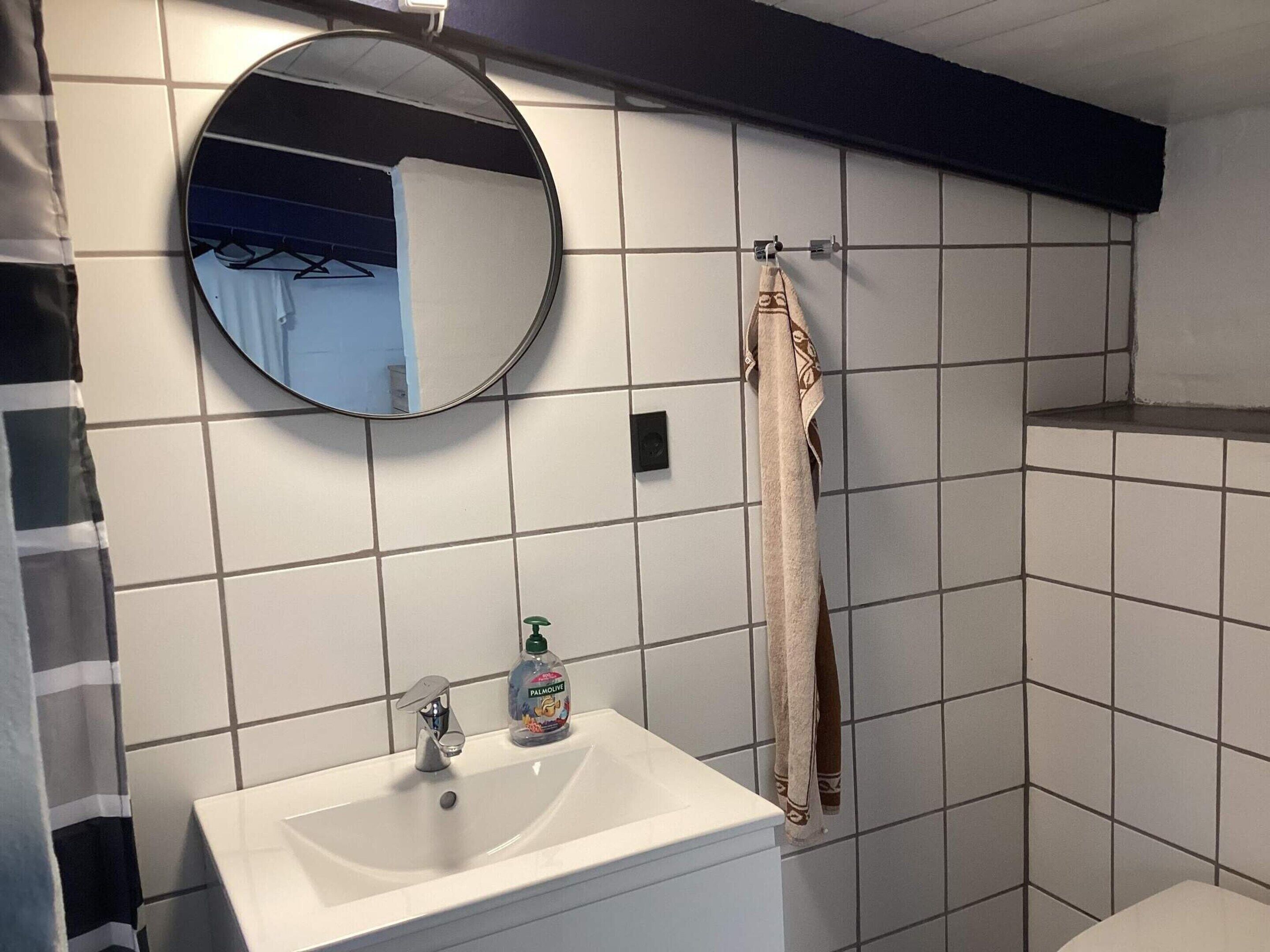 Un bany amb un lavabo blanc, un mirall rodó, un penjador de tovalloles i una zona de dutxa.