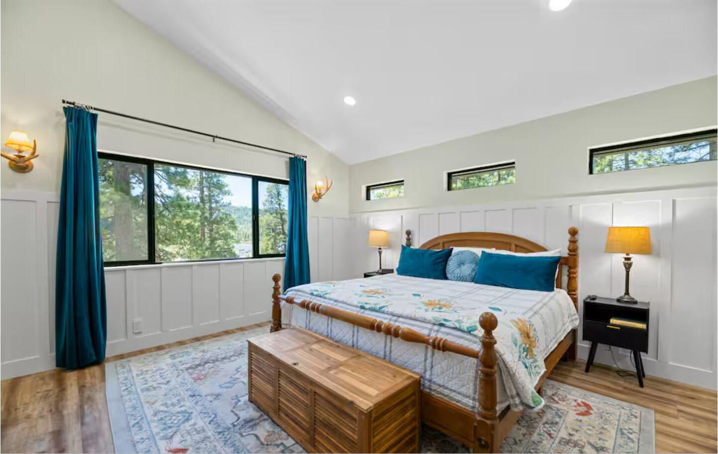 A bedroom with a large bed, two bedside tables, a wooden bench, and a view of trees through the windows.