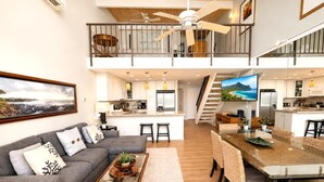 A spacious living area with a grey sofa, a coffee table, and a dining table with chairs. A kitchen is visible in the background with a TV mounted above the counter. - Napili Point | Oceanfront 2 BR | Water Front | Car Incl. w/ 6+ Nights | NAP-C19 by KBM (Lahaina)