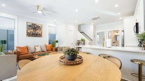 Un intérieur moderne avec une table à manger ronde en bois, un canapé avec des coussins orange, un ventilateur de plafond et un coin cuisine visible à travers une porte ouverte.